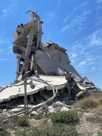 The image shows a severely damaged or destroyed building in Az-Zahra, Palestinian Territories. The building is constructed primarily of concrete, now reduced to rubble and fractured slabs. Twisted metal rebar is visible within the debris. Some sections of the structure remain vaguely recognizable as supporting beams and walls. The sky is blue with wispy clouds. Vegetation grows near the base of the destroyed building.