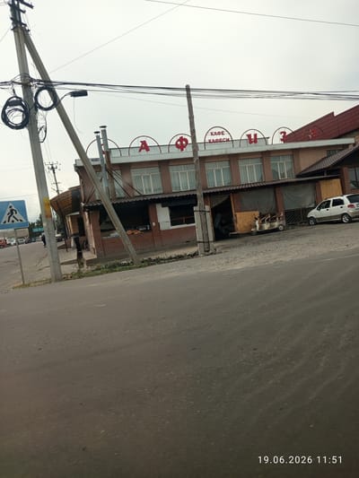 The image shows a building with a sign that reads "КАФЕ КАФЕСИ" (Cafe Cafesi) in red lettering. The building appears to be a commercial establishment, possibly a restaurant or cafe, with large windows and an outdoor seating area. There are several vehicles parked in front of the building, including a white car and a three-wheeled scooter. A pedestrian crossing sign is visible on the left side of the image, indicating the presence of a road and traffic. The overall environment suggests an urban or semi-urban setting. The timestamp "19.06.2026 11:51" is superimposed on the lower right corner of the image.