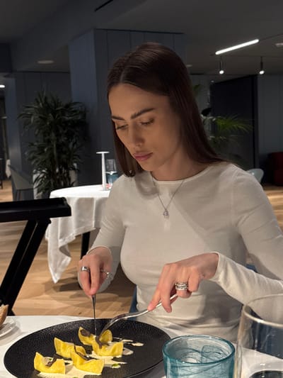 A woman with long, dark brown hair, wearing a light-colored, long-sleeved top and a silver-colored pendant necklace, is seated at a table in a restaurant located in Torbole, Italy. She is looking downwards, actively cutting food on a black, textured plate with a silver fork in her left hand and a silver knife in her right hand. The plate holds several large, yellow pasta shapes, covered in a cream-colored sauce and topped with dark, thinly sliced material, consistent with truffle. Rings are visible on multiple fingers of both hands. A blue-green patterned glass is present on the table to her right, and a piece of bread on a small white plate is visible to her left. The restaurant interior is dimly lit, characterized by dark walls, a wooden floor, and several tall potted green plants, resembling palms, in the background. Other dining tables, set with white tablecloths and glassware, are visible further into the room. Modern linear light fixtures are mounted on the ceiling.