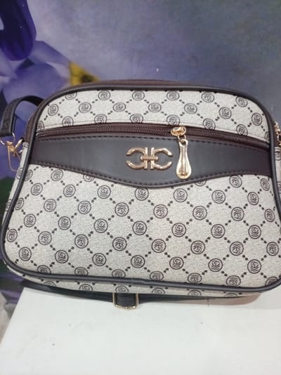 This is a close-up, eye-level shot of a beige and brown patterned handbag. The handbag has a repeating geometric pattern of interlocking circles with a stylized symbol inside, set against a diamond-shaped grid. There is a dark brown band across the front of the bag, above a zippered compartment. A gold-colored zipper pull in the shape of a teardrop hangs from the main zipper. Centered on the brown band is a gold-colored logo, appearing to be two stylized letters intertwined. The handbag has dark brown trim and a dark brown strap attachment on the left side, with a gold-colored buckle visible at the bottom. The background is a blurred image of a large purple flower with a yellow center, suggesting a decorative or floral backdrop. The handbag rests on a white surface, possibly a table or shelf. The lighting appears to be artificial, and there are no clear indicators of time of day or weather. The image does not contain any visible text. The overall impression is that of a product photograph, likely for sale or display.