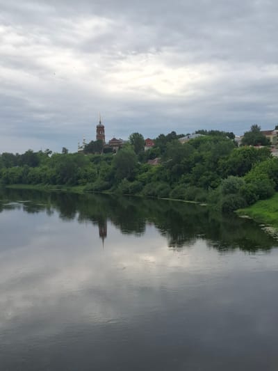 The image captures a scenic view of the Iren River in Kungur, Russia, with a prominent church steeple reflected in the calm waters. The foreground is dominated by the dark, reflective surface of the river, with subtle ripples distorting the mirrored image of the sky and the opposite bank. The middle ground features a lush, green embankment densely covered with trees and vegetation. Beyond the trees, on a slight rise, stands a traditional Russian church with a tall, brick bell tower topped with a golden spire. Several other buildings, likely residential, are scattered among the trees on the hillside. The sky above is overcast with soft, diffused light, casting a muted tone over the entire landscape. The overall atmosphere is serene and tranquil, characteristic of a riverside town on a cloudy day.