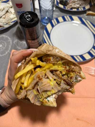 An overhead photograph depicts a dining table scene in Γεωπονικά, Greece. A fair-skinned hand, exhibiting manicured fingernails with light polish and subtle gold accents, holds a large, partially consumed gyros or souvlaki wrapped in brown paper. The food item is densely packed with sliced meat (likely pork), numerous golden french fries, a yellow mustard-like sauce, and a white tzatziki-like sauce; a distinct bite has been taken from its lower right side. The hand wears a dark-colored wristband or smartwatch band. The food is positioned over an orange rectangular placemat, which rests on a grey patterned tablecloth. In the background, from left to right, are a partially visible plate containing food remains (possibly fish bones), a white plastic container labeled "ΓΑΛΑ" with a red logo, an open black aluminum beverage can, a ridged blue drinking glass, and an empty round plate with a blue and yellow striped rim. A transparent plastic fork is visible on the orange placemat to the right of the gyros.