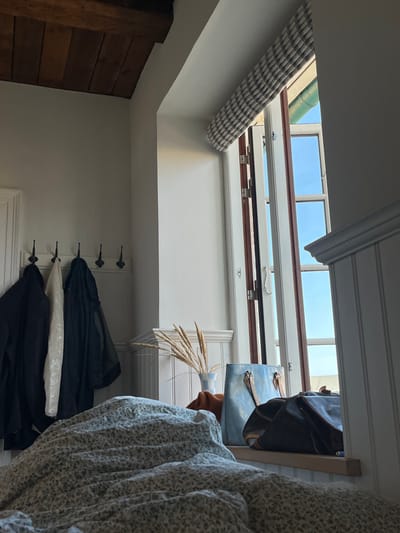 This image depicts an interior room, likely a bedroom, located in Skagen, Denmark. The view is from a low vantage point, presumably from a bed, with a light-colored duvet patterned with small dark specks or flora covering the lower portion of the frame.

To the left, a white wall features a coat rack mounted at mid-height, equipped with five dark, double-pronged hooks. Three garments hang from the hooks: a black jacket, a white textured garment, and a dark, possibly translucent, jacket. Above, dark wooden ceiling beams are visible across the top-left quadrant of the frame.

To the right, a large, multi-paned window is open inwards, revealing a clear blue sky and a distant light-colored building or structure beyond. The window has white frames and dark brown sashes. A light-colored Roman blind with a grid pattern is partially rolled up at the top of the window frame. Below the window, a white wainscoting paneling extends along the wall. On the wide windowsill, a light-colored vase contains several stalks of dried ornamental grass. Adjacent to the vase, two bags are placed: a light blue tote bag with tan handles and a dark brown, possibly leather, satchel or messenger bag.