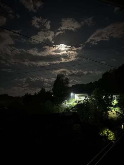 The image depicts a night scene in Ogradna, Bulgaria. The moon is visible through clouds, illuminating them. Utility wires are stretched across the sky. A building, likely a residence, is visible amidst trees on the right. A bright light source is present near the building. The foreground is predominantly dark.