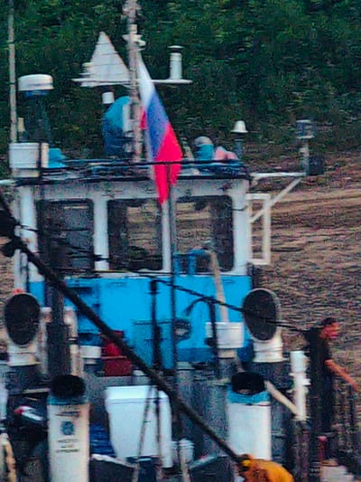 The image shows a close-up view of a boat with a Russian flag flying. The boat appears to be docked or anchored, with trees and foliage visible in the background, suggesting a natural setting. The boat has a blue and white cabin with windows. Various pieces of equipment, possibly related to maritime operations, are visible on the deck, including pipes, cylindrical structures, and a black circular object.
A person in a dark shirt is standing on the right side of the boat, with their right arm extended, potentially interacting with something out of frame or signaling. Another person, partially visible at the bottom of the frame, is wearing an orange jacket. The scene appears to be during the day, with diffused lighting. The overall impression is one of a functional boat in a rural or natural environment. No text is visible in the image.