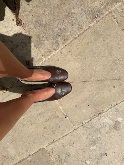 A photograph depicts a first-person perspective of a person standing on a paved surface, likely in Saint-Victor-la-Coste, France, evidenced by the style of paving. The subject's legs and feet are visible, with the person wearing dark brown leather slipper-style shoes adorned with blue gem-like embellishments on the vamps. The paving stones are light grey and irregular, forming a pattern of interlocking rectangles, and are marked by slight weathering. To the left of the frame, a dark, possibly metal, object is visible, potentially a part of a bollard or other street furniture. The scene is bathed in sunlight, creating shadows along the pavement.