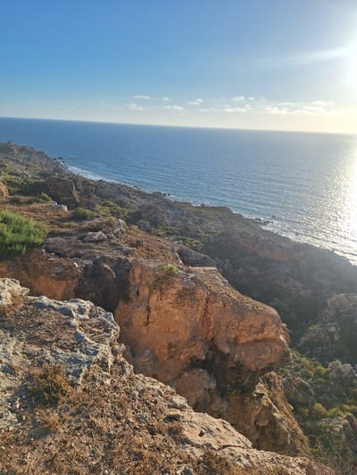 The media file displays a coastal cliffside view overlooking the Mediterranean Sea near Ahrara, Morocco. The scene is dominated by rugged, rocky terrain with patches of dry brush and sparse green vegetation clinging to the slopes. The cliffs are steep and uneven, with layers of rock formations visible.

The sea is a deep blue, with sunlight glinting off its surface, suggesting a bright and sunny day. A hazy horizon line separates the sea from a pale sky, with a few wispy clouds present. The sun, positioned in the upper left quadrant of the image, casts a bright glare, indicating it is likely late morning or early afternoon. The overall impression is one of a natural, untamed coastal landscape. There are no people or man-made objects visible, emphasizing the wild and remote character of the location. The image evokes a sense of peace and natural beauty, characteristic of a coastal environment.
