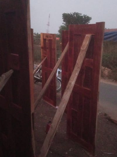 Multiple dark reddish-brown paneled wooden doors are observed standing upright in an outdoor environment in Ebiya, Nigeria, under an overcast sky. Each door is individually propped by one or more light-colored wooden planks leaning diagonally against its surface. Approximately five doors are visible in various stages of presentation across the foreground and mid-ground. Behind the central doors, a white motorcycle is partially discernible. The immediate ground consists of uneven, reddish-brown soil with scattered light-colored debris. A small red cylindrical object rests on the ground near the base of a central door. To the right of the doors, a dark asphalt road is visible. In the distant background, green foliage and a red and white lattice communication tower are present against the light grey sky. No individuals are visible.