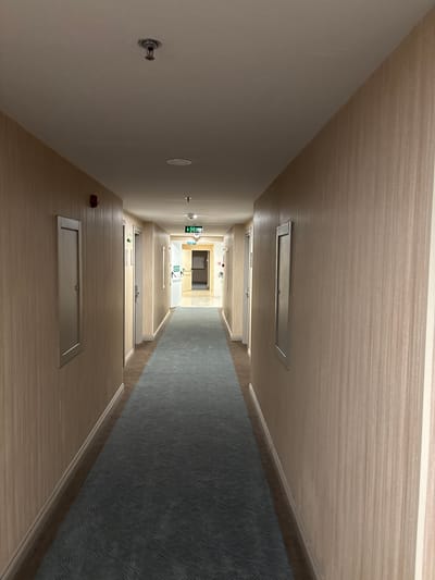 The image depicts a long, empty interior hallway, likely within a modern hotel or apartment building in Selçuk, Turkey. The perspective looks down the corridor, which recedes into the distance, ending in a brightly lit area.
The floor of the hallway consists of a light-colored, wood-like laminate or tile bordering a central strip of dark blue or grey patterned carpet. The left wall is a plain light beige, while the right wall and the ceiling are covered in a consistent light beige material with a subtle horizontal wood-grain texture. White baseboards line the bottom of both walls.
Numerous light-colored doors are visible along both sides of the hallway. Rectangular, silver-colored access panels are embedded in the ceiling. The corridor is illuminated by artificial light sources, including recessed ceiling lights.
On the right wall, in the mid-distance, a green emergency exit sign clearly displays the word "EXIT" with an arrow pointing to the right. Below the English text, there is text in a different, indiscernible foreign script, likely Turkish (ÇIKIŞ). Closer to the foreground on the right wall, a small, red, bell-shaped fire alarm pull station or sensor is mounted, alongside an ornate, decorative metal fixture.
The hallway is devoid of people or activity, conveying a sense of quiet and solitude. The modern design and clean appearance suggest a well-maintained or recently constructed facility.