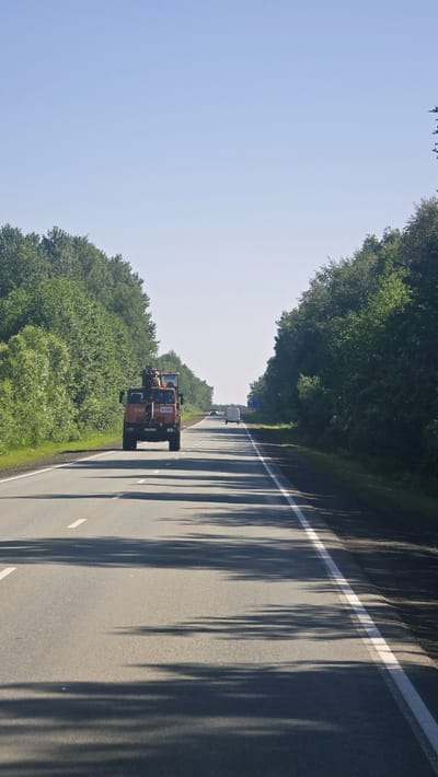 An orange truck with a crane attachment is driving down a two-lane road lined with dense green trees on both sides. The sky is clear and blue. There are other vehicles visible in the distance, including a white van. The road has white lane markings and the sun is casting shadows from the trees onto the asphalt. The scene appears to be on a sunny day in a rural or suburban area.
