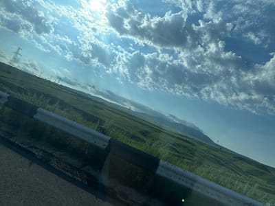 The image shows a landscape with a road in the foreground, bordered by a guardrail. Beyond the road, rolling green hills stretch into the distance, dotted with power lines and transmission towers. The sky is blue with scattered clouds, and sunlight streams through, casting rays. The scene appears to be a rural area, possibly in or near Solak, Armenia, given the context. There are no people or visible actions taking place in the image. The environment is natural, with vegetation covering the hills and a paved road. The guardrail is made of metal and has black and white reflective markings. The overall impression is of a scenic, undeveloped area.