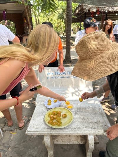 This is an outdoor photograph taken at daytime in Tân Thạch, Vietnam. The scene features a group of people gathered around a stone table. A blonde woman in a pink tank top is taking a photo with her pink phone. Another person wearing a large straw hat is partially obscuring the view. On the table, there is a light green plate of Vietnamese savory pancakes called "Banh Khot" served with a dipping sauce and a small plate of the same type of pancakes. Some individuals are taking the snacks from the plate. The table has text engraved on it, which reads "DNTN TRƯƠNG TH." underneath which is a phone number "ĐT: 0275.2473504 - 0961.111049". The surrounding environment includes greenery, with trees and foliage visible. Some shelter-like structures are present in the background with red lanterns.