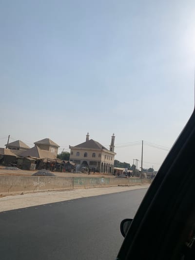 The image depicts a roadside scene in Gora, Nigeria, under clear, sunny skies. In the immediate foreground, a freshly paved, dark asphalt road is visible. A low, grey concrete barrier with "30+000" painted in white lettering separates the road from the adjacent terrain. The right side of the frame is partially obscured by a dark vehicle window frame and side mirror.
Beyond the barrier, a diverse collection of structures and activity unfolds. Prominently featured in the center is a light-colored, two-story building, likely a mosque, characterized by a central domed roof, arched windows on the upper level, and an arcade of arches on the ground floor. Two slender minarets with conical tops flank the main structure.
To the left of the mosque, multiple single-story buildings with light-colored walls and hip roofs are visible, interspersed with several informal, possibly makeshift, structures constructed from corrugated metal or similar materials. Piles of grey aggregate (gravel) are scattered on the ground.
Numerous individuals are present, clustered around the buildings and piles of material. Some appear to be standing or walking. One area in front of the mosque shows a plume of light-colored smoke or dust rising from ground level. Further to the right, a power pole with multiple overhead lines extends vertically. The background consists of a clear, light blue sky, with the sun's glare visible in the upper right quadrant.