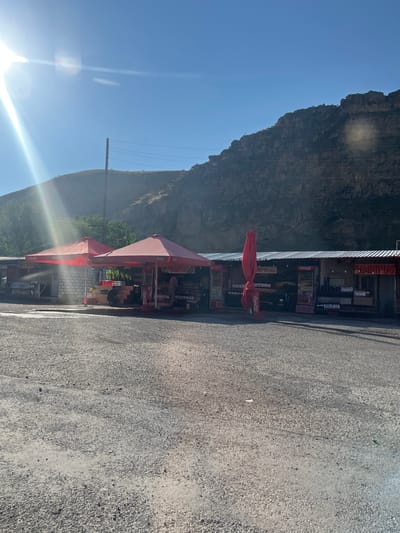 This is an eye-level, full-shot photograph of a roadside market in Zangakatun, Armenia, under a clear blue sky. The sun is bright, casting a strong glare and lens flare on the left side of the image. In the foreground, there is a paved area with small gravel. Behind it, a row of market stalls with red awnings are visible. These stalls appear to be selling various goods, including drinks displayed in refrigerators and on shelves. One stall has a Coca-Cola branded refrigerator. A tall, folded red umbrella stands near one of the stalls. Behind the stalls, a green tree line is present, and further in the background, a large, rocky mountain dominates the scene. A wooden utility pole with wires runs across the middle ground. Two individuals are partially visible within the market stalls, appearing to be interacting with the goods or customers. The overall impression is a typical roadside scene in a rural or mountainous area.