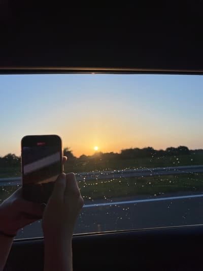 In Shchyrets, Ukraine, at sunset, a person is seen from the chest up, holding a smartphone in their hands, capturing the view from a car window. The phone's screen displays a recording indicator and a diagonal white line, possibly representing a road or reflection. Outside the window, a hazy sunset paints the sky in gradients of blue and orange, with the sun a bright orb just above the tree line. The foreground includes the dark interior of the car, framing the window. Tiny water droplets or specks are scattered across the car window, distorting the view slightly. The overall scene suggests a moment of travel and observation, with the person documenting the picturesque sunset.