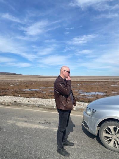 A bald, light-skinned man wearing a brown leather jacket, black pants, and black boots stands on the asphalt shoulder of a road in Kliuchiki, Russia. He has sunglasses on and his left hand is near his face. The front of a silver car is partially visible to his right. Beyond the shoulder of the road, there's a muddy, barren field enclosed by a fence. The sky is blue with scattered white clouds.