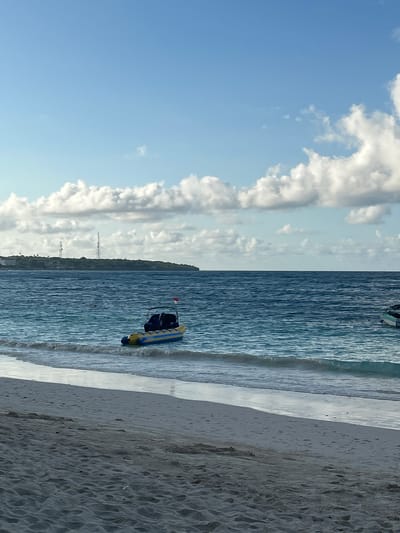 An eye-level, wide-angle shot captures a tranquil beach scene in A R A, Indonesia. The foreground is dominated by a sandy beach, with the gentle waves of the ocean lapping at its edge. In the middle ground, a yellow and blue inflatable banana boat is anchored in the calm, blue waters, equipped with a dark canopy and what appears to be two seats. Further in the distance, a lush green island with two tall communication towers is visible. The sky above is a clear blue, dotted with scattered white clouds. The lighting suggests it is daytime, likely mid-morning or late afternoon. The overall atmosphere is serene and peaceful.