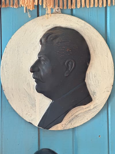 The image shows a round bas-relief portrait of Joseph Stalin, in profile, rendered in dark metal against a light-colored, weathered background. The portrait is mounted on a blue wooden surface, which appears to be a door or wall panel, with the grain of the wood clearly visible. Above the portrait, a fringe of peach-colored thread or yarn hangs from the top edge of the frame. The overall impression is of a historical artifact displayed in a rustic setting. The location provided, Бигила, Russia, is a village in the Kirov Oblast.