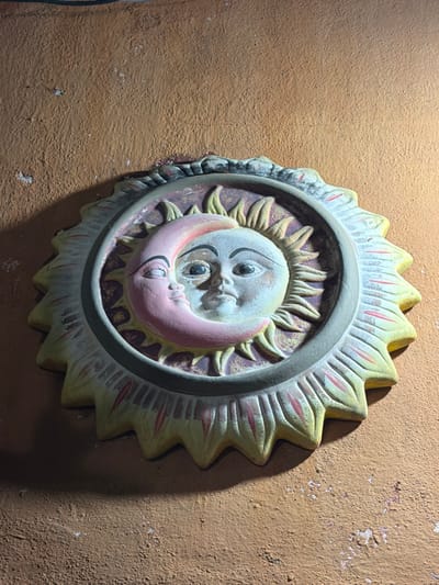 A circular, decorative wall hanging depicts the sun and moon. The sun's face is pale gray, surrounded by stylized rays painted pale yellow with red accents. The moon, rendered in pink, nestles below. Both faces have closed eyes, creating a peaceful expression. A gray ring encircles the central sun and moon image. The entire piece is attached to a textured orange wall with some chipped sections, and there is a dark cable visible to the left.