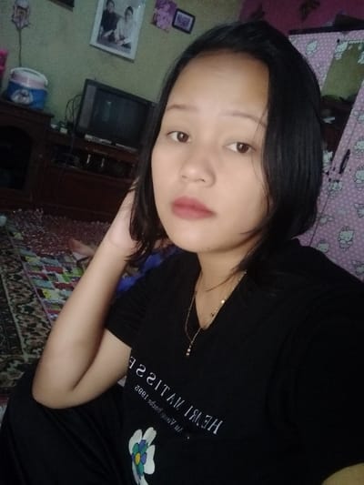 A young Asian woman poses for a selfie. She has dark hair, and wears a black t-shirt with white text and a flower design. A gold necklace adorns her neck. Her makeup is natural. The background includes a TV, a rug with an ornate pattern, and a wall adorned with pictures and a Hello Kitty themed curtain. The overall ambiance suggests a casual, home setting.