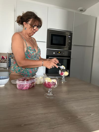 The image shows a woman wearing a floral swimsuit in a modern kitchen. She is in the process of serving ice cream into two stemmed glasses. In front of her on the counter are two containers, one with pink ice cream and the other with what appears to be a white substance. The kitchen features white cabinets, a stainless steel microwave, and a wooden countertop. The scene appears to be in the city of Saint-Just-Saint-Rambert, France.