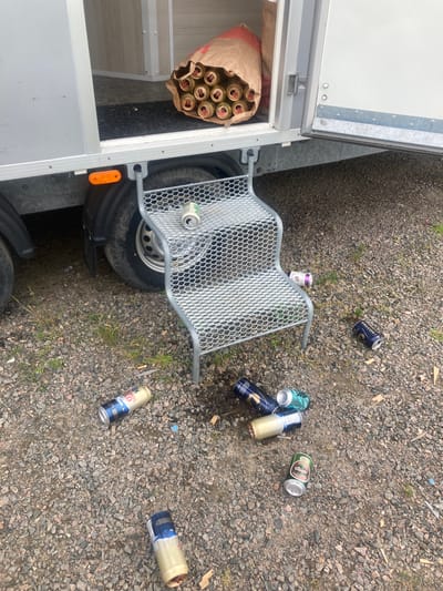 This image captures an outdoor scene in Vessigebro, Sweden, presenting the aftermath of a beverage consumption event adjacent to a parked trailer. A silver-colored utility trailer with an open side door is positioned on the left side of the frame. Inside the trailer, a brown paper bag contains approximately ten metallic gold-colored beverage cans with red pull tabs, appearing full or unopened. A two-tiered, grey, mesh-grate metal step is placed at the trailer's open doorway. An empty light green aluminum can rests on the top tier of this step. The ground consists of light brown and grey gravel or crushed stone, with sparse patches of green grass and weeds emerging through it. Scattered across this gravel are at least nine empty, discarded aluminum beverage cans of various brands, including "Lands Wild," "Falcon," and "Tuborg Classic"; several are dented or partially crushed. Small liquid spills are visible on the gravel near the base of the steps, along with cigarette butts and other minor debris. No individuals are present in the scene.