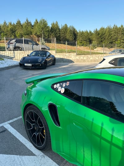This media file depicts a bright green sports car, likely a Porsche, parked outdoors. The car is the primary subject, with its sleek design and vibrant color dominating the frame. A black sports car is parked behind it. The scene appears to be an outdoor parking area, possibly a lot or a road, with a concrete barrier and a metal fence visible to the left. Beyond the fence, a dense line of trees forms a backdrop, suggesting a somewhat rural or natural setting. The ground is paved with asphalt, marked with white lines. The sun is casting distinct shadows, indicating it is likely daytime, possibly late afternoon given the angle of the light and the warmth of the colors. The sky is clear and blue. There are several stickers on the rear window of the green car, but they are too small to read. The license plate of the black car is visible and reads "CB 0911 HM". The overall impression is one of parked luxury vehicles in an open-air environment.