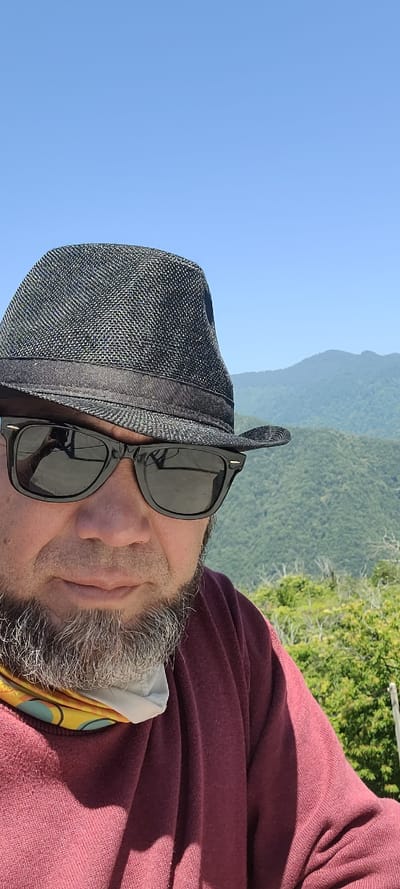 A medium shot is captured of a person wearing a black fedora and black sunglasses, facing the camera. The subject has graying facial hair and is wearing a maroon shirt with a colorful bandana around their neck. The background features a lush, green, mountainous landscape under a clear blue sky. This scene is located in Milisi, Georgia.