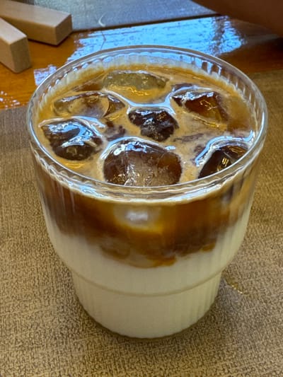 The image shows a close-up, high-angle view of an iced coffee drink in a clear glass cup. The coffee is layered, with a lighter cream or milk mixture on top of a darker coffee base, and ice cubes are visible within the drink. The glass has a ribbed texture on the lower half. The drink is placed on a textured surface that appears to be a light brown or beige tablecloth or placemat. To the left of the cup, a wooden surface is visible with two small wooden blocks. A hint of a person's hand or arm can be seen in the upper left corner of the image. The lighting suggests it is daytime, possibly indoors given the consistent lighting and the tablecloth. The scene appears to be a casual setting, possibly a cafe or a home. There is no visible text in the image.