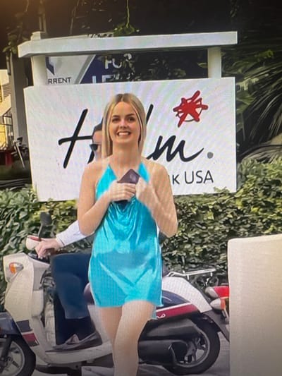 A young woman with blonde hair and a wide smile is pictured standing next to a large white sign with a red star-like logo and black lettering. She is wearing a vibrant turquoise dress. The sign has text that appears to read "M" and "KUSA". To the right of the woman, a portion of a white scooter with red and blue accents is visible. The background consists of lush green foliage, suggesting an outdoor setting, possibly during the day. The woman's expression conveys joy and excitement. The sign's design and the scooter suggest a potentially tropical or resort-like location, though no explicit text confirms this. The orientation of the image is rotated 90 degrees counter-clockwise.