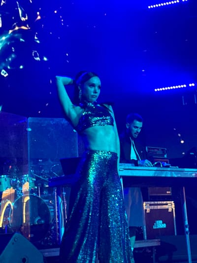 This is a photograph of a woman performing on stage, illuminated by blue lights. She is wearing a sparkling sequined outfit consisting of a halter-neck crop top and wide-leg pants. Her hair is pulled back in a ponytail, and she is wearing large earrings. Her left arm is raised behind her head. In the background, a man is visible behind a DJ setup, and drum equipment can be seen to the left. The overall environment appears to be a concert or performance venue. This scene is not located in Akhalubani, Georgia.