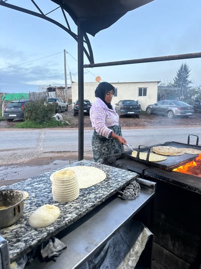 The image captures an outdoor scene depicting a woman cooking flatbreads on a large griddle. The image is oriented vertically, with the top of the scene on the left and the bottom on the right.

The central figure is a woman, dressed in a dark head covering (possibly a hijab or scarf), a light purple or lavender-colored knitted cardigan with a patterned design, and a dark patterned skirt. She is actively engaged in cooking, holding a long-handled spatula or flipper, which she uses to tend to several round flatbreads sizzling on a large, dark metal griddle. Visible flames emanate from the left side of the griddle, indicating a direct heat source, possibly wood-fired or a strong gas burner.

Adjacent to the cooking area, to the right of the woman, is a countertop with a speckled gray and white pattern, resembling granite or marble. On this counter, several pieces of dough are prepared for cooking: a large, flat round piece, a smaller oval-shaped piece, and a distinctive cylindrical piece with horizontal ridges, suggesting a specific preparation technique for layered flatbreads. Further to the right on the counter, there are metal containers or troughs; one contains a crumpled gray cloth, and another is partially covered with a dark material.

The background reveals an unpaved or dirt road, along which several vehicles are parked or visible in the distance, including a dark sedan, another dark car, and lighter-colored vehicles. Simple, light-colored buildings, one with a window, are visible beyond the road. Sparse trees, possibly conifers, and some low-lying bushes dot the landscape. The sky is overcast and uniformly gray, suggesting a cloudy day, rather than bright sunshine. A dark, metallic, open-frame structure, likely part of an awning or a makeshift stall, partially frames the upper left corner of the scene. An orange satellite dish or antenna is mounted on a pole in the middle ground, and a green fence or netting can be seen in the far distance.

The scene portrays a roadside or outdoor food stall specializing in freshly made flatbreads in what appears to be a semi-urban or rural setting. The woman appears focused on her task. No discernible text is visible in the image.