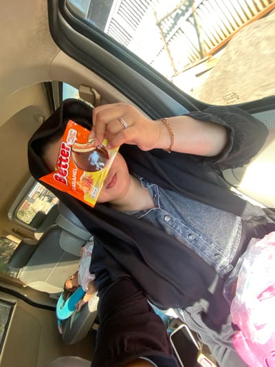 A person wearing a black hijab and a denim shirt is sitting in the passenger seat of a vehicle. The person is holding an orange wrapper with the word "Better" and "Caramel" printed on it, partially covering their face. A child is visible sleeping in the back seat on a blue neck pillow. The environment appears to be an urban area, possibly the city of Ngrambe, Indonesia, given the sunlight and the corrugated metal structure visible through the window. The car's interior is primarily beige and grey.