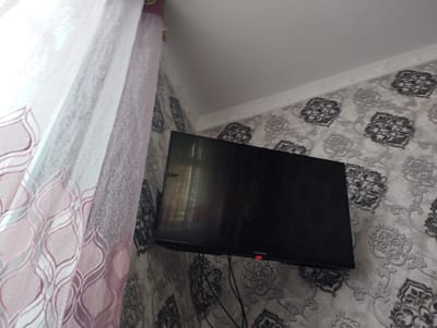 The image displays an interior room, likely in Mingchinar, Uzbekistan, characterized by decorative elements. A black, wall-mounted flat-screen television is the central subject, positioned towards the right side of the frame. The TV screen is dark, showing reflections of the room, and a small red indicator light is visible on its bottom right bezel, alongside the "SAMSUNG" logo. Several dark cables hang from the bottom of the television.

To the left, a set of curtains partially obscures a window, through which diffused bright daylight enters the room, suggesting it is daytime. The inner curtain is sheer and white or very light pink, while the outer curtain features a distinctive, repeating wavy or leaf-like pattern in a slightly darker pink hue.

The walls are adorned with wallpaper featuring an ornate, grey and white/light grey damask or floral pattern. A strip of white crown molding or coving runs along the top edge of the wallpaper, meeting a plain white ceiling. No people are visible, and no particular activity is taking place; the scene is static.
