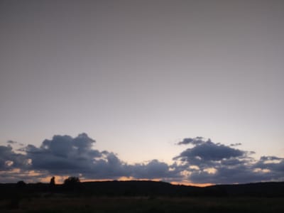 This is a photograph of a landscape at dusk in Cherevky, Ukraine. The sky is a soft gradient of pale gray at the top, transitioning to a light pink and orange hue near the horizon. Dark, textured clouds are scattered across the middle ground, some appearing heavy and shadowed, others catching the fading light. Below the sky, a silhouetted, hilly terrain is visible, with dark shapes of trees and vegetation dotting the landscape. In the far distance, the faint outline of a man-made structure or a prominent tree can be discerned against the horizon. The foreground consists of a darker, blurred area that suggests fields or uneven ground. The overall mood is tranquil and serene, characteristic of twilight.
