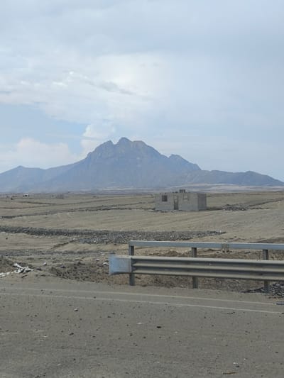 The image depicts a landscape in Huanchaco, Peru, under an overcast sky. In the background, a large, multi-peaked mountain dominates the horizon. In the midground, a flat, arid plain stretches out, marked by rudimentary stone walls and what appears to be the incomplete construction of a small structure, possibly a house, made of grey bricks. The foreground is dominated by a stretch of asphalt road, bordered by a metal guardrail. The overall impression is one of a sparsely populated, undeveloped area with a rugged, natural backdrop.