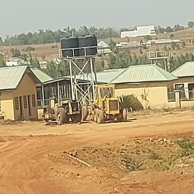 The scene, located in Haifam, Nigeria, displays an undeveloped or construction area under a clear, bright sky. In the foreground, reddish-brown dirt ground is visible with tire tracks and disturbed earth. A white pipe segment lies partially exposed on the ground. The midground features three single-story buildings with light yellow facades and green corrugated metal roofs. The leftmost building has visible windows. Centrally positioned, a tall metal framework tower supports two black cylindrical water tanks. Two large yellow heavy construction vehicles, resembling motor graders, are parked on the dirt, one partially obscured by the water tower's base and the other to its right. A smaller, darker structure with an open-air or veranda appearance is situated between the left and right buildings, behind the water tower. The background reveals rolling hills covered with sparse green vegetation and numerous scattered residential or light industrial structures extending into the distance. No individuals, active actions, or interactions are discernible.