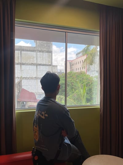 A person is seated on a red bench facing a large window, with their back to the camera, looking out at a view of buildings and trees. The room has yellow-green walls and is partially framed by dark brown curtains. The person is wearing a long-sleeved shirt with Japanese writing and graphics on the back, and dark pants. A bag with a smiley face charm is visible. Outside the window, a weathered, multi-story concrete building with visible rebar and a rusted red roof is prominent. Beyond that, lush green foliage and palm trees are visible, along with another building with rows of windows. The sky is partly cloudy. The scene appears to be a view from a room in Dumai City, Indonesia.