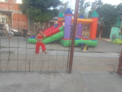 An outdoor scene displays children near an inflatable bounce house. A young girl in a red outfit stands in front of a wire fence. Other children are seen in the background, some near a car. The bounce house is colorful with red, green, purple, and orange sections, set up on a concrete street. Houses and trees are in the background.