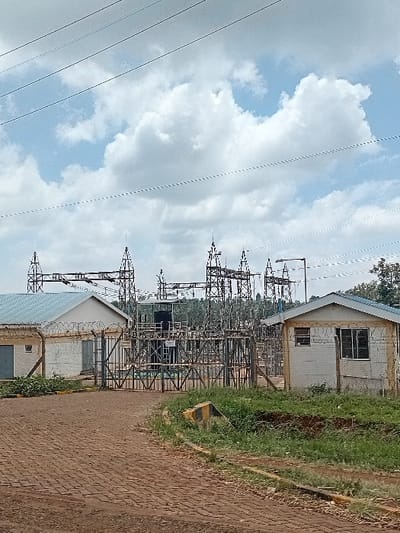 The image depicts an electrical substation in Mururi, Kenya, under a partially cloudy sky. In the foreground, a brick-paved access road leads towards a large, closed metal gate with wire mesh and barbed wire at its top center. To the left of the gate, a single-story white building with a light blue corrugated metal roof, multiple doors, and windows is partially visible, enclosed by a wire fence topped with barbed wire. To the right of the gate, another smaller, single-story white building with a light blue corrugated metal roof and barred windows is visible, also surrounded by a wire fence with barbed wire. Patches of green grass and exposed soil flank the paved road, with a yellow and black striped concrete barrier on the right side. Beyond the gate, the substation complex features numerous metal lattice towers supporting electrical equipment, including power lines, insulators, and transformers. Multiple overhead power lines cross the upper portion of the frame. No individuals or vehicles are present.