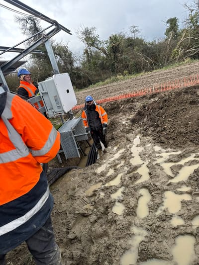 An outdoor work site at Baldurgan Hill, Ireland, displays three male workers in varying positions on muddy terrain under an overcast sky. In the foreground, the back of a worker in an orange high-visibility jacket with grey reflective stripes and dark trousers is visible, facing towards the left side of the frame. To the mid-left, a worker in an orange high-visibility jacket, blue hard hat, and a beige head covering stands next to a metallic solar panel mounting structure. This structure supports a white rectangular electrical enclosure with warning labels and a grey perforated cable tray, from which black corrugated conduits emerge into a muddy excavation. A third worker, wearing an orange high-visibility jacket, dark trousers, dark sunglasses or safety glasses, and a blue hard hat, stands within this muddy, water-filled trench to the right. The ground is heavily disturbed earth, characterized by deep mud, numerous puddles, and tire or boot tracks. Orange plastic mesh safety fencing lies partially on the ground in the mid-background. The background consists of a line of deciduous trees and bushes with bare branches.