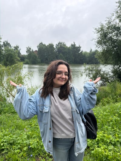 A young woman with dark, wavy hair and glasses stands in front of a body of water, possibly a lake or river, in Nikulskoye, Russia. She is wearing a light blue denim jacket over a striped long-sleeved shirt and blue jeans. Her arms are outstretched, palms up, in a gesture that could be interpreted as questioning or welcoming. A black backpack is slung over her right shoulder. The background consists of lush green vegetation and trees, with a slightly overcast sky above. The overall impression is that of a casual outdoor setting.