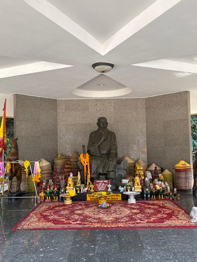 A statue of a seated monk is centered in a niche with a granite-like wall. Offerings line the shelf above and below the statue, including miniature figures, floral garlands, and small structures. A patterned red rug extends in front of the niche. Below the rug, two wooden horses are positioned. A colorful flag is visible on the far left. The ceiling is white with geometric patterns and recessed lighting.
