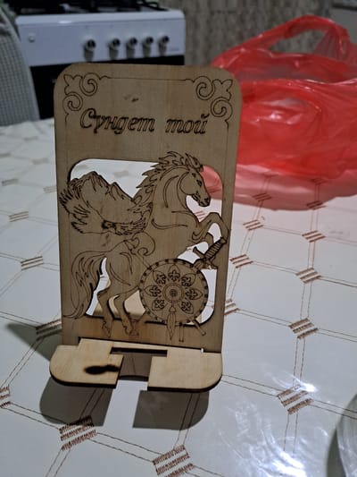 A wooden phone stand is displayed on a white tablecloth with a geometric pattern. The stand is carved with the image of a horse, a sword, a shield, a mountain, and an eagle. Above the image, the Cyrillic text "Сүндет той" is inscribed. To the right of the stand, a crumpled red plastic bag is visible. In the background, a white appliance with four knobs is partially visible, likely a stove or oven. The scene appears to be indoors. The city of Ынтымақ ауылдық округі, Kazakhstan is not depicted in this image.