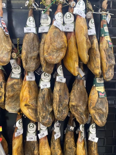 The image depicts a retail display of various cured hams, likely jamón ibérico, hanging in rows. The hams are brown and have a textured, cured appearance. Many of the hams are adorned with labels, some of which display prices, such as "139" and "180," and branding like "MONTE CERVERO" and "LEGADO IBÉRICO." The background consists of a dark tiled wall. The scene suggests a food market or a specialty store in Bel Air, Spain, where these hams are presented for sale. No people are visible in the image.