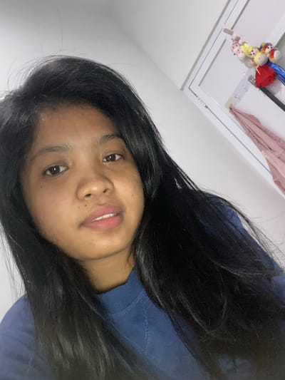 A young person with dark hair and brown eyes is angled towards the camera. They are wearing a blue shirt. The background includes a white wall, a section of a white ceiling with a light fixture, and what appears to be a few small plush toys attached to the ceiling near the light. The location is inferred, but there's no visual confirmation in the image.