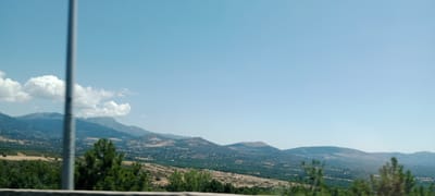 The image depicts a wide, panoramic view of a mountainous landscape under a clear blue sky with scattered white clouds. In the foreground, a blurred vertical pole, likely a traffic or utility pole, partially obscures the view. The midground reveals rolling hills covered in vegetation, transitioning into rugged, distant mountains. The slopes are dotted with trees and appear to have some areas of scrubland or dry grass. The overall impression is a rural or natural setting, possibly taken from a moving vehicle given the slight blur. The location is identified as Guadarrama, Spain, suggesting this is a view of the Guadarrama mountain range.