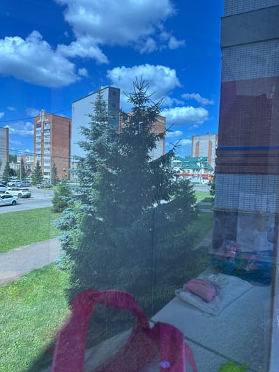 The image, taken from a window, shows a residential area in Mariinsky, Russia, on a sunny day with a bright blue sky and scattered white clouds. In the foreground, a large evergreen tree partially obscures the view. Beyond the tree, a paved walkway and a patch of green grass are visible, with several parked cars on the street. Residential buildings, including multi-story apartment complexes, are present in the background, some with brick facades and others with light-colored siding. A building with a green roof and arched windows is also visible in the distance. The window itself shows some reflections, including a pink object in the bottom left corner and a white surface with some items on it to the right.