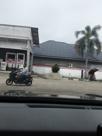 A man in a striped shirt and denim shorts is riding a black scooter on a paved surface. He is wearing glasses and has short dark hair. The scooter is moving towards the left of the frame. In the background, there is a building with a white and grey facade and a dark tiled roof. Two pink and white gas pumps are visible on the left side of the building, with a sign reading "JALUR BBM SUBSIDI". A palm tree stands to the right of the building, and a pile of sand is located in front of the building. A rusty tricycle-like structure is parked near the palm tree. The sky is overcast. The image appears to have been taken from inside a vehicle, as the lower portion of the frame shows the dashboard and windshield. The scene is consistent with a gas station in Meunasahlhok, Indonesia.