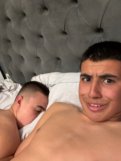 The image displays two male subjects in a bedroom, identified as Kanchevo, Bulgaria. In the foreground, on the right, a light-skinned male with short dark hair and a light mustache is visible from the chest up, looking directly towards the viewer with a slight smile. He is bare-chested. Adjacent to him, on the left side of the frame, another light-skinned male with short dark hair is positioned, appearing to be asleep with his face turned away from the camera, resting on a light-colored pillow. Both individuals are in a bed with light-colored bedding. A grey, tufted upholstered headboard is prominent in the background.